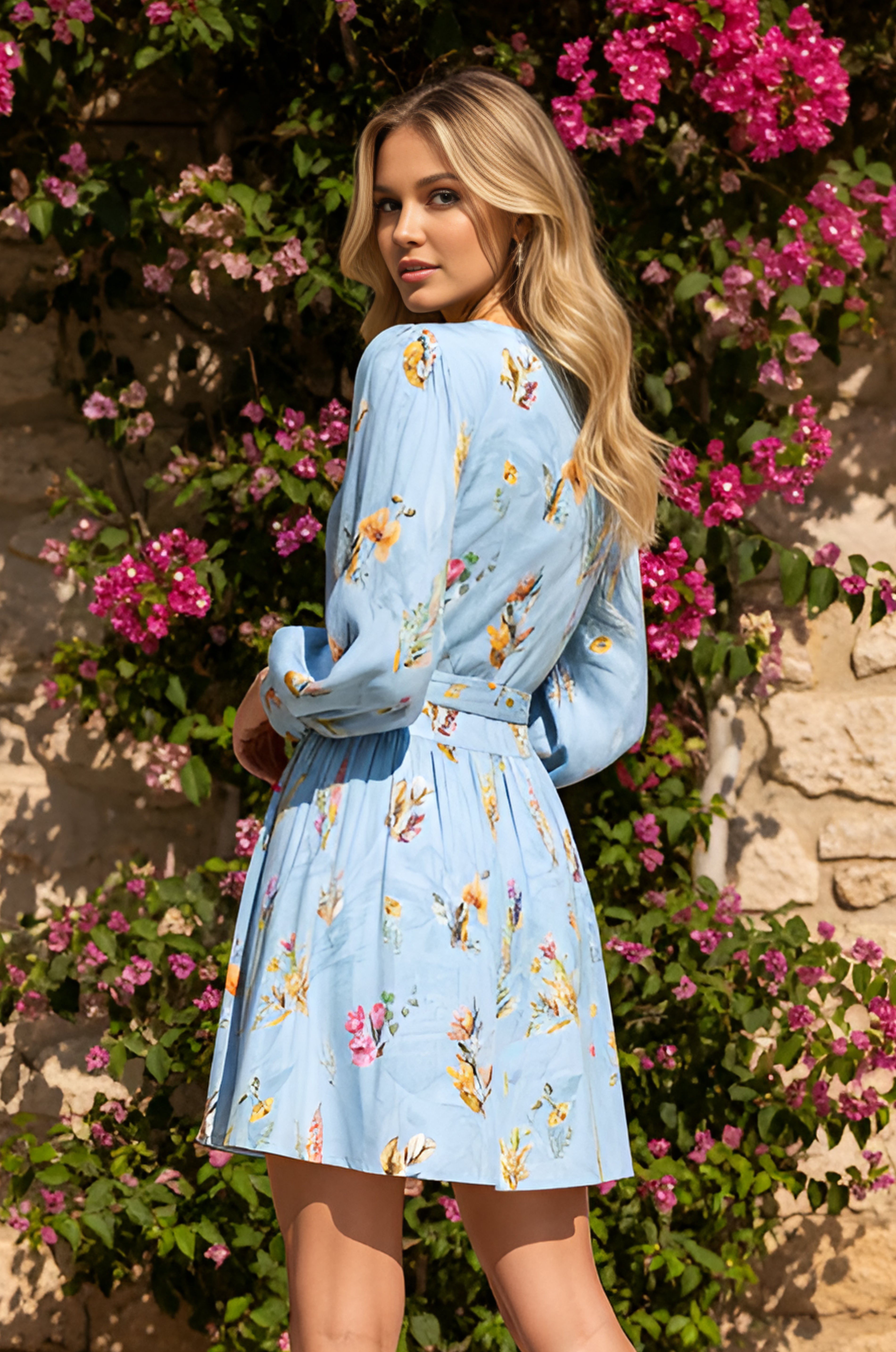 A woman with long blonde hair, wearing the Sky Blue Short Casual Dress: Paige, stands outdoors by a stone wall and blooming pink flowers, looking over her shoulder.